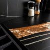 Noir St Laurent Porcelain worktop surface detail with black background and warm coppery veining beside countertop items.