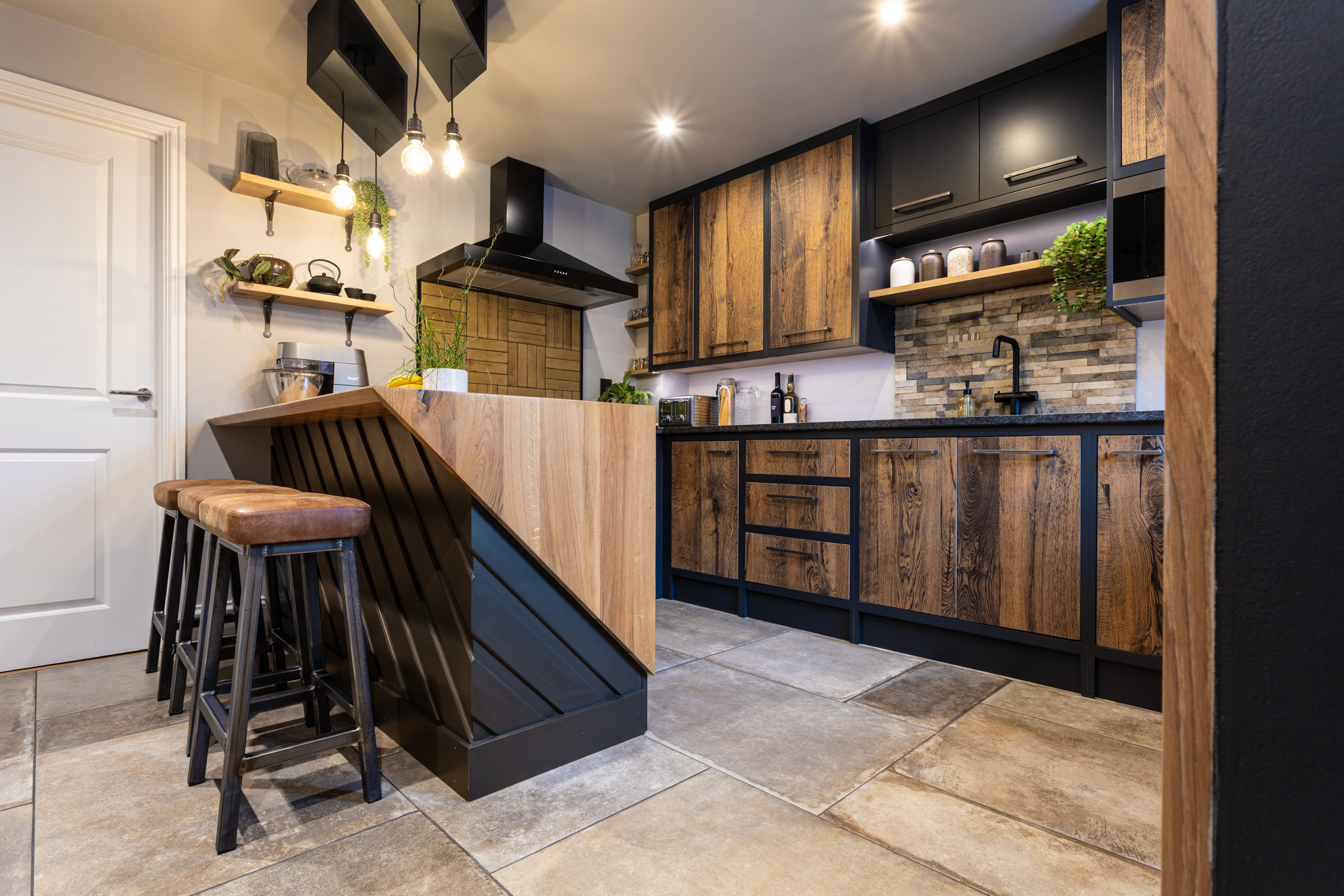 Wide angle kitchen view with Steel Grey leather Granite worktops and an island breakfast bar, showing rustic wood cabinetry, bar stools and stone effect flooring.