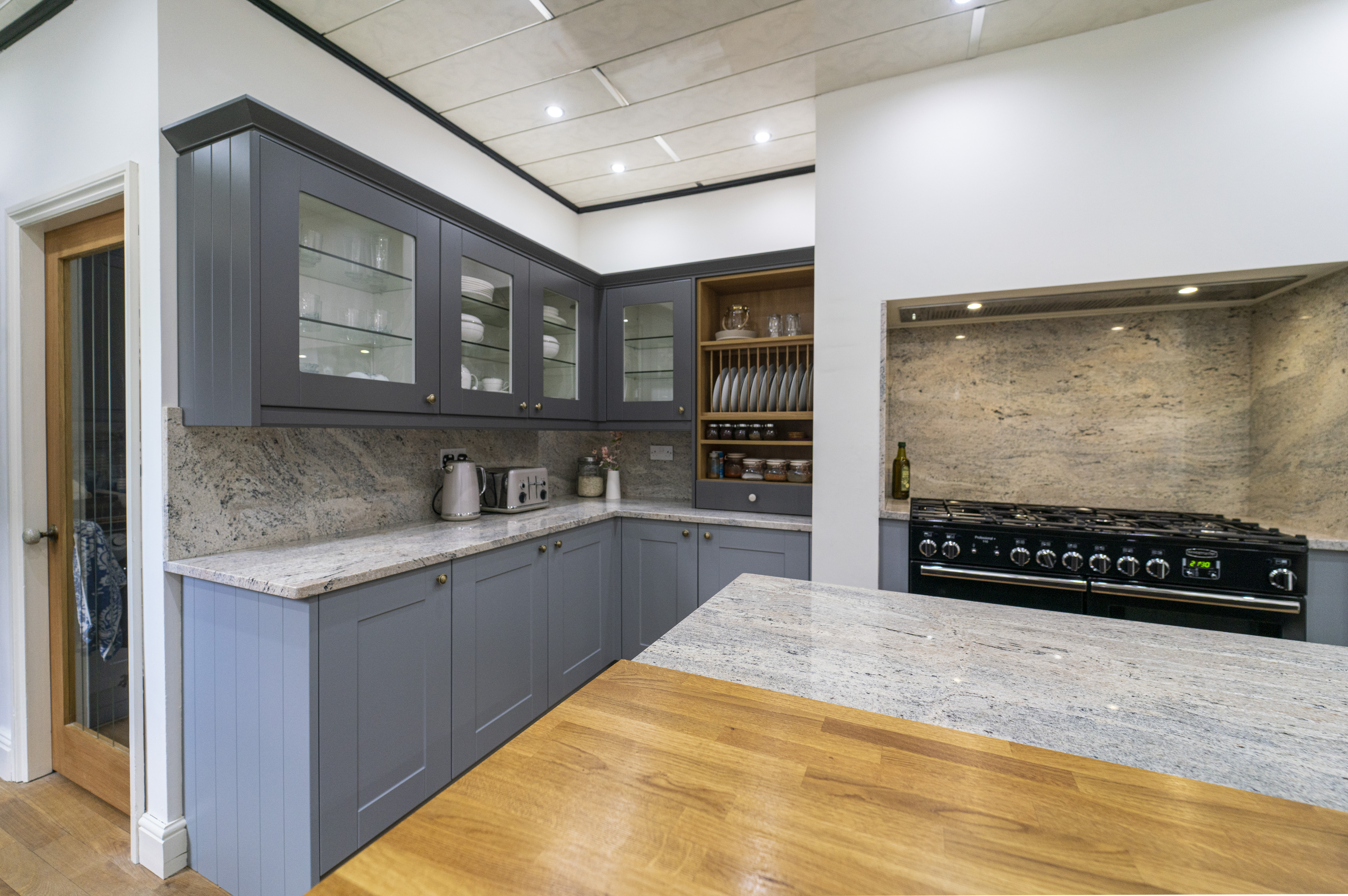 Wide kitchen view showing Ivory Fantasy Granite worktops along the main run and peninsula, with blue cabinetry, timber worktop section and a range cooker alcove.