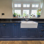 Front view of a blue kitchen sink run with Absolute Black Granite worktops, showing a white farmhouse sink, tap and window plants above the worktop.