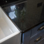 Close up of an Absolute Black Granite worktop corner with a polished finish, showing the edge detail above blue cabinetry next to a white sink.