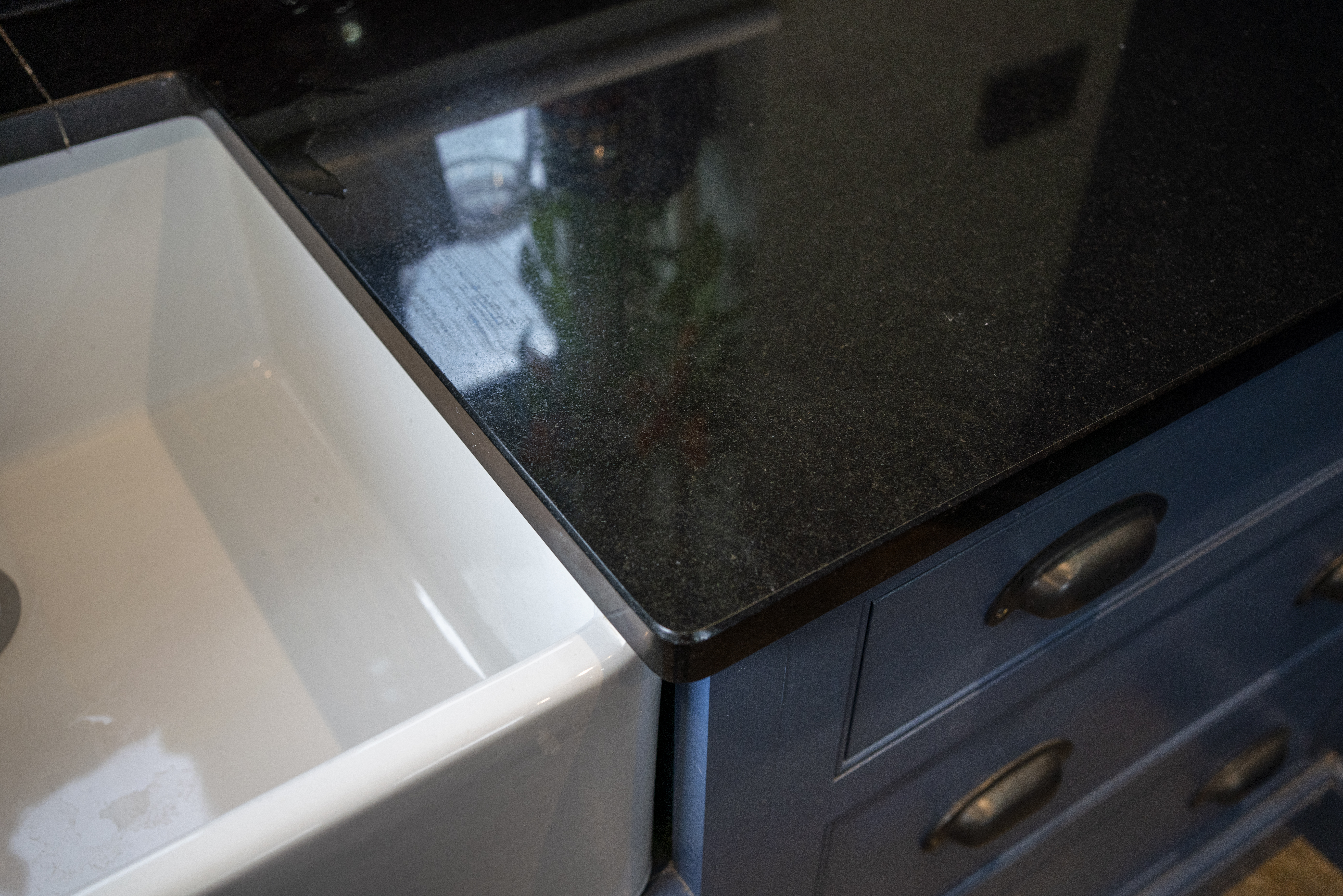 Close up of an Absolute Black Granite worktop corner with a polished finish, showing the edge detail above blue cabinetry next to a white sink.
