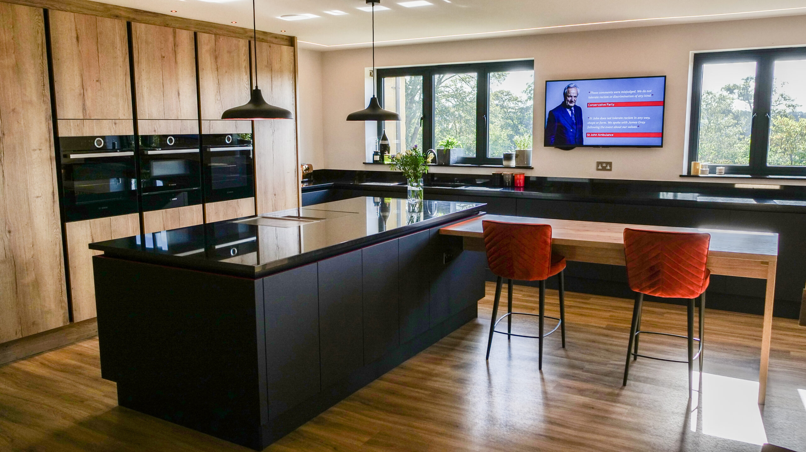 Open plan kitchen view with an Absolute Black Granite island and breakfast bar seating, showing pendant lights, sink area and large windows.