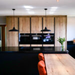 Kitchen view showing an Absolute Black Granite island worktop with breakfast bar and seating, with timber wall units and built in ovens in the background.