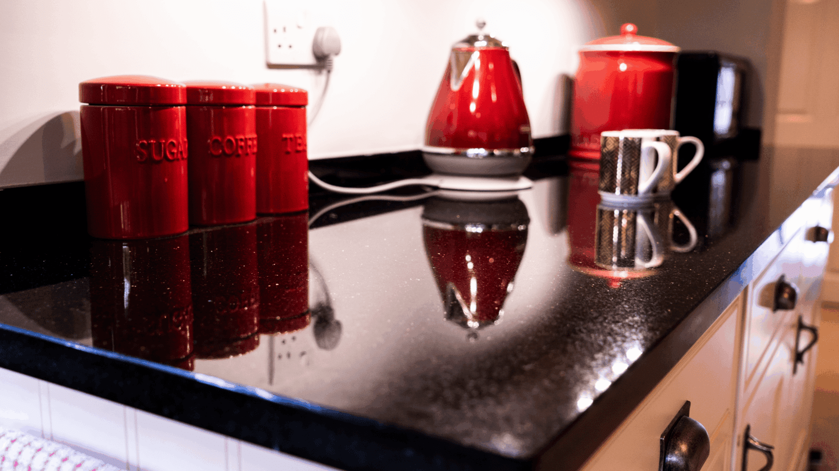 Close up of Star Galaxy Granite worktop with a polished reflective finish, showing sparkling flecks and red kitchen accessories reflected on the surface.