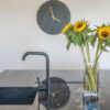 Close up of a sink area in Cosmic Black Granite with a black tap, showing the polished reflective surface and sunflowers in a vase on the worktop.