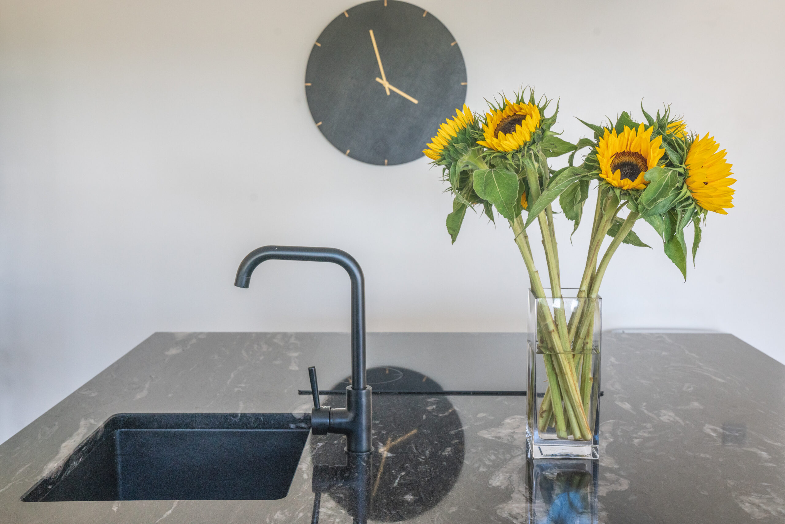 Close up of a sink area in Cosmic Black Granite with a black tap, showing the polished reflective surface and sunflowers in a vase on the worktop.