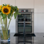 Close up of a Cosmic Black Granite worktop with a black tap and polished reflective surface, styled with a vase of sunflowers in a modern kitchen.