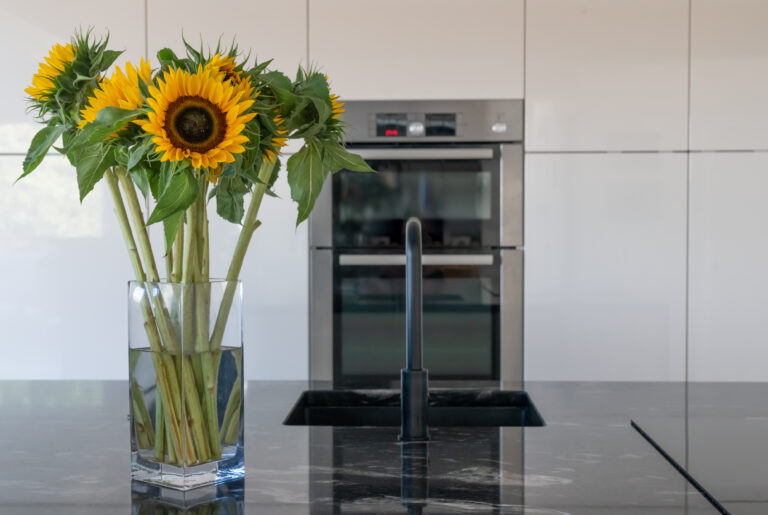 Close up of a Cosmic Black Granite worktop with a black tap and polished reflective surface, styled with a vase of sunflowers in a modern kitchen.