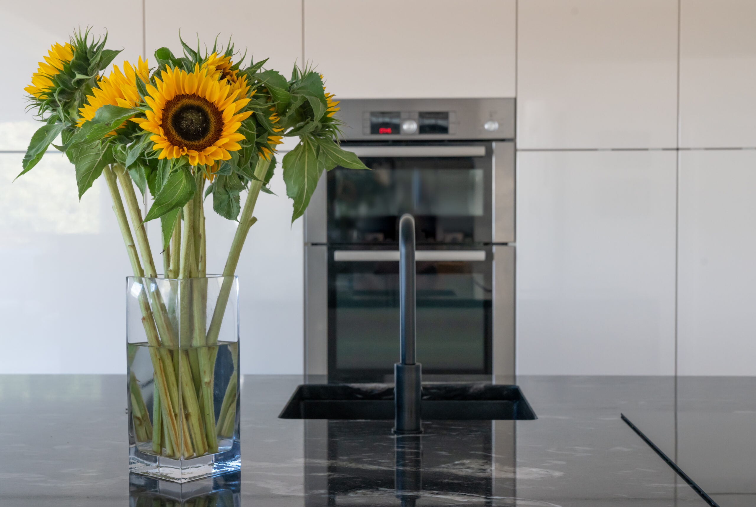 Close up of a Cosmic Black Granite worktop with a black tap and polished reflective surface, styled with a vase of sunflowers in a modern kitchen.