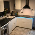 Open plan kitchen angle showing Blue Pearl Granite worktops on white cabinetry, with under cabinet lighting and the speckled Granite surface along the run.