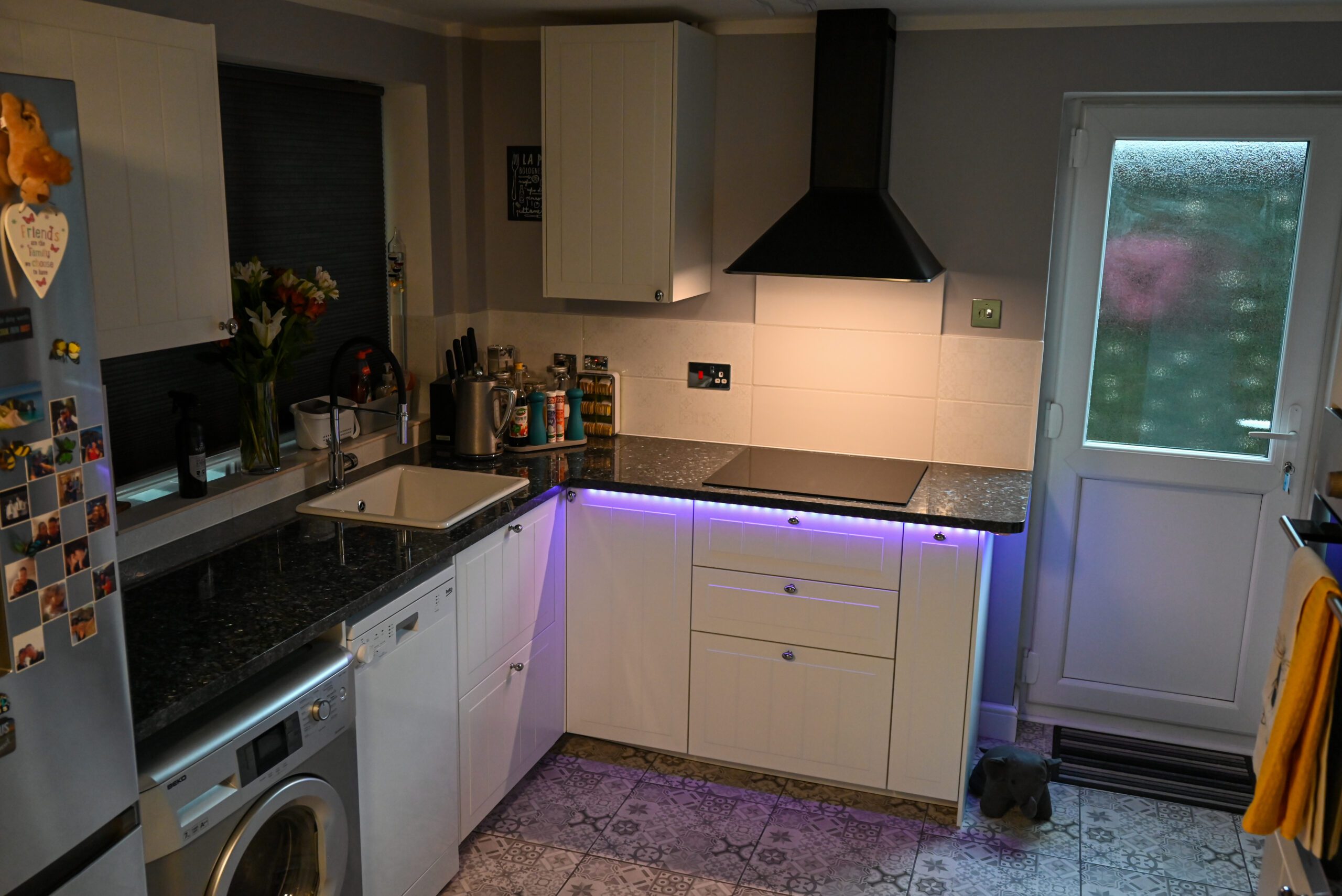 Wide kitchen view with Blue Pearl Granite worktops and under cabinet lighting, showing a corner run with white units and a dark speckled Granite surface.