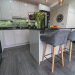 Wide kitchen view showing an Absolute Black Granite island worktop with breakfast bar seating, with grey stools, white cabinetry and a green tiled splashback.