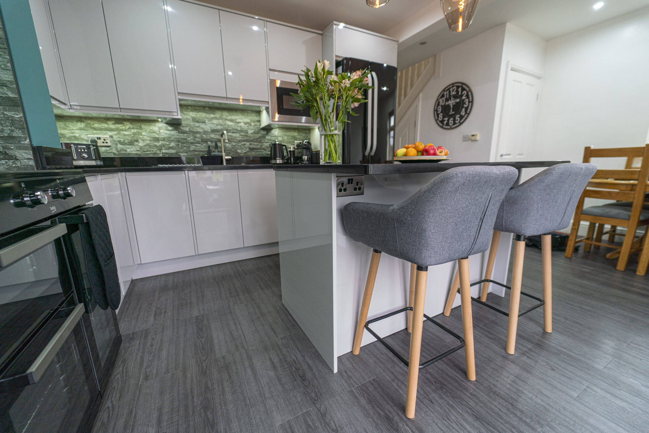 Wide kitchen view showing an Absolute Black Granite island worktop with breakfast bar seating, with grey stools, white cabinetry and a green tiled splashback.