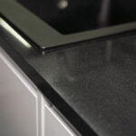 Close up of an Absolute Black Granite worktop corner with a polished finish, showing the edge detail beside an inset hob in a modern kitchen.
