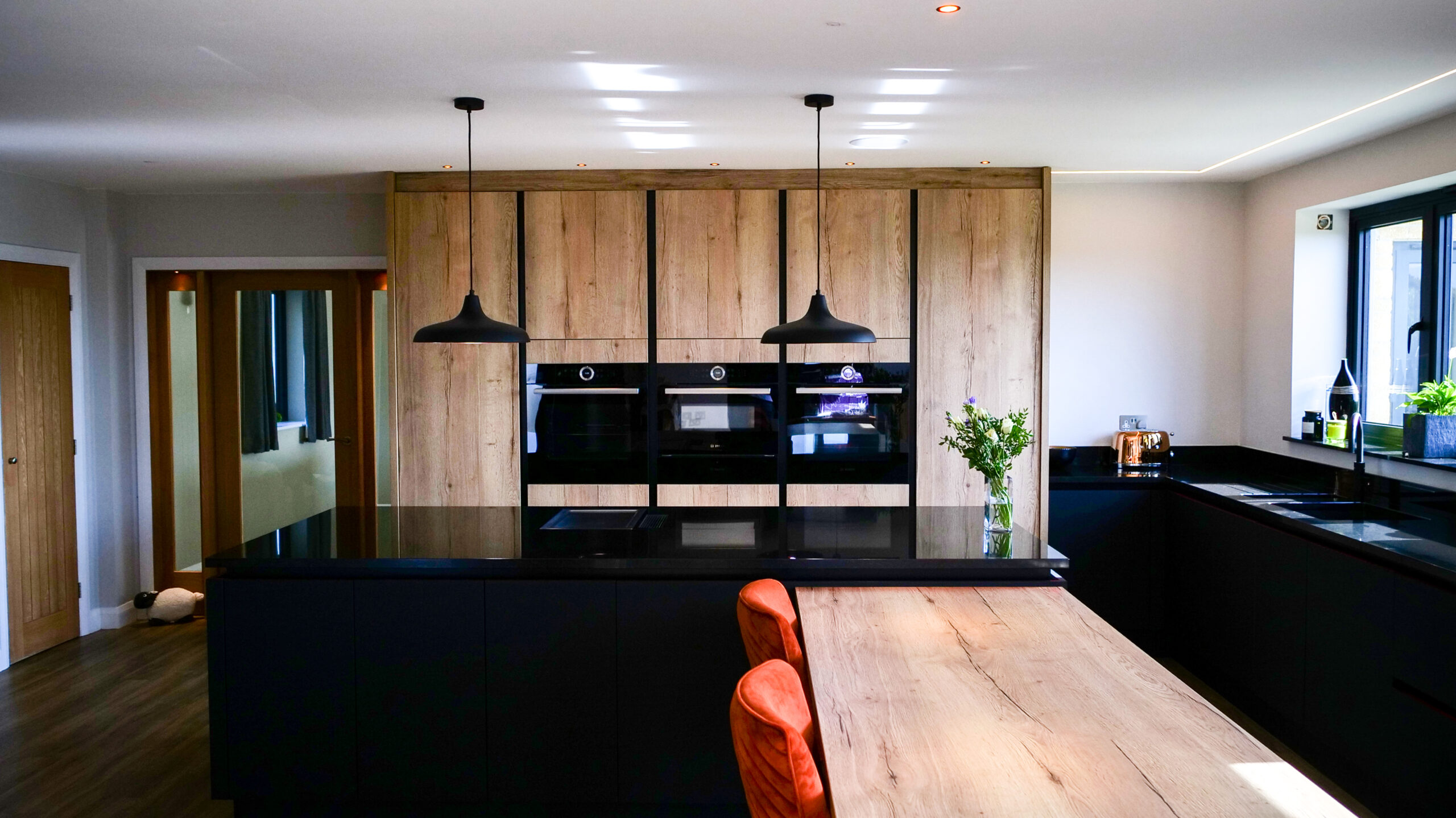 Wide kitchen view with an Absolute Black Granite island worktop and breakfast bar seating, showing timber wall units, pendant lighting and open plan layout.